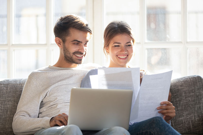 A smiling couple sits on a couch, looking at documents together. The man has a laptop on his lap, and both people appear happy and focused. Bright window light fills the room behind them.