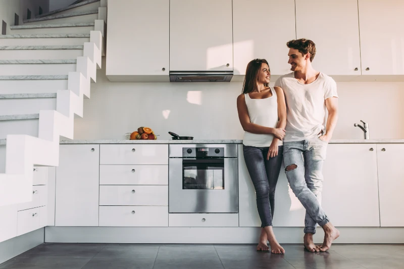 A young couple, both casually dressed in white tops and jeans, stands barefoot in a modern white kitchen, smiling at each other while leaning against the counter near a staircase.