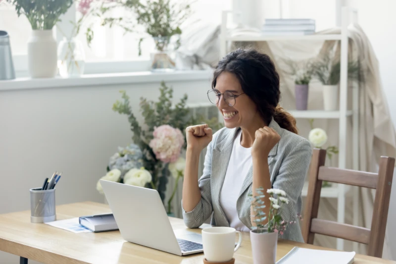 A woman sits at a desk with a laptop, smiling and raising her fists in excitement. The room has plants and flowers, and sunlight streams through a window in the background.